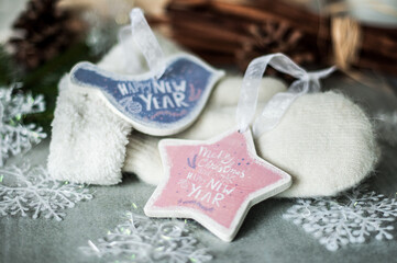 Wooden Christmas toys of bird and star, lying on warm mittens with artificial snowflakes and snow, fir branches, pine corns and bundles with cinnamon sticks on a gray ceramic tiles closeup.