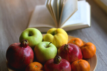 Wooden bowl filled with various seasonal fruit and open book on a table. Selective focus.