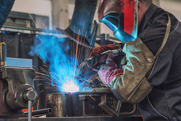 A heavy industrial worker in a factory works with metal on angle grinders while hot sparks are...