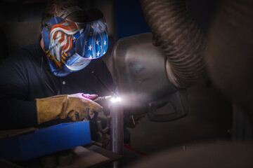 A heavy industrial worker in a factory works with metal on angle grinders while hot sparks are...