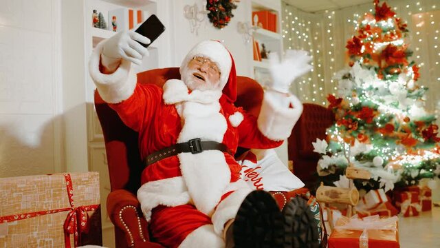 Santa Claus, holding a smartphone in his hands, smiles and congratulates people on Christmas and New Year through video communication