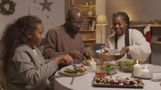 Tracking shot of African-American family of three sitting at dinner table and enjoying Christmas dishes while celebrating together in cozy living room