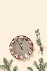 Winter festive table setting concept, Christmas plate with clock hands show twelve or midnight, sparkle balls beige pink color, knife, fork, fir tree branches. New Year holiday food concept