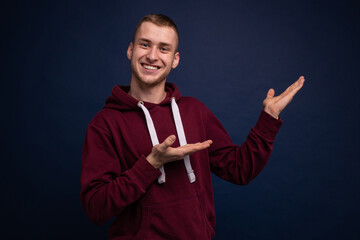 Young man in red hoodie and jeans posing on a blue background, smiling and showing palms to the side