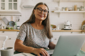 Pretty middle-aged woman with grey hair working on her laptop sitting against kitchen background, playing online games or watching movie, having happy face expression. People, technology, leisure time