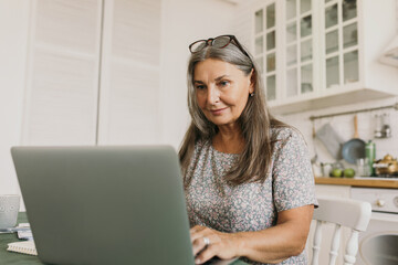 Picture of beautiful, elegant 60-years-old woman with glasses on head typing on notebook keypad...