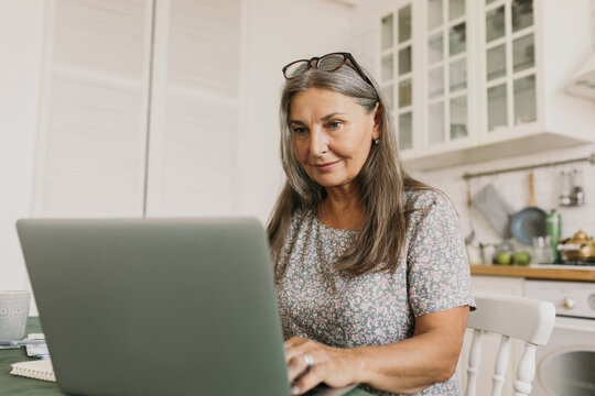 Picture of beautiful, elegant 60-years-old woman with glasses on head typing on notebook keypad browsing internet using wireless connection from home, sitting in white Scandinavian style kitchen