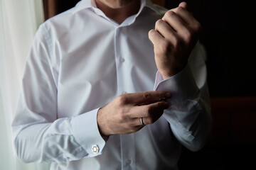 Man buttoning cuff link of luxury white shirt sleeve, copy space. Groom fixes cufflinks standing...