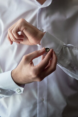 Man buttoning cuff link of luxury white shirt sleeve, copy space. Groom fixes cufflinks standing near window, close up