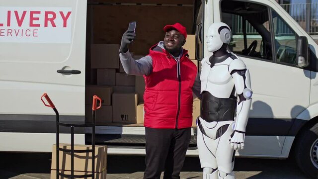 Delivery man standing in front of delivery van with robot, making selfie. African american courier delivering parcel box to recipient.