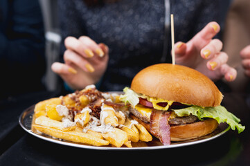 Close-up gourmet bacon burger with fries on table and unrecognizable female hands on background.