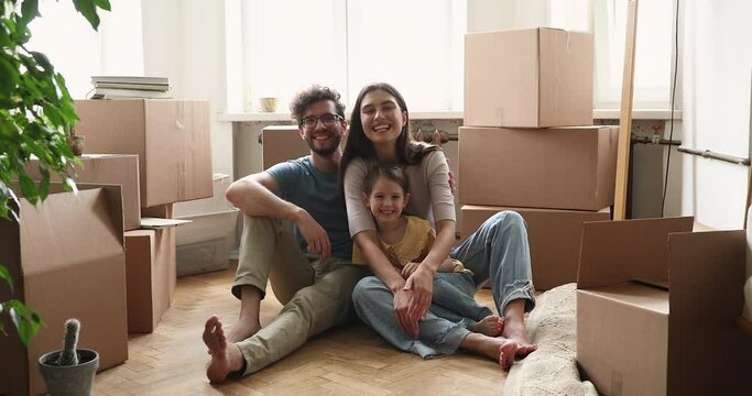 Family portrait loving young parents little girl pose on floor at moving day sit among big boxes cuddle look at camera with glad smiles. Happy mom dad child receive mortgage loan do renovation at home