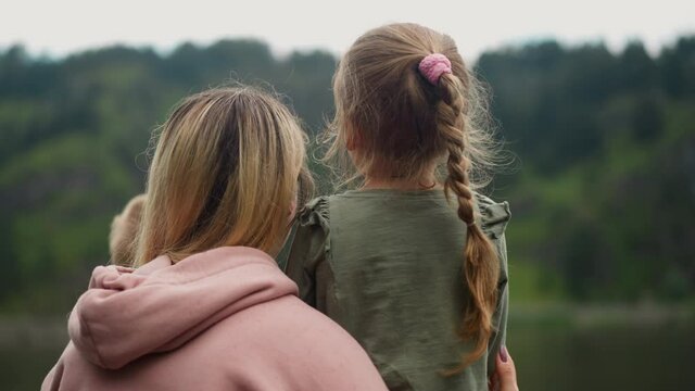 Happy woman hugs little children talking to daughter while looking at hills covered with forest on cloudy rainy cool day backside view