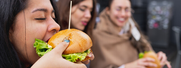 Horizontal banner or header Side view young woman biting a burger. Females sitting at table in restaurant.