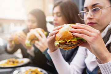 SIde view three young women sitting at table eating burgers. Close-up holding hamburger and bite it.