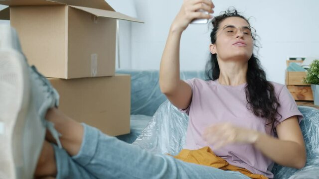 Cheerful young woman is chatting on video call in new house after relocation pointing at boxes talking and laughing sharing positive emotions.