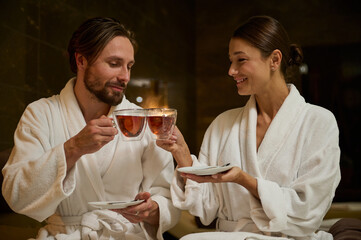 Happy newlywed, charming middle aged married Caucasian couple in white bathrobes holding cups with herbal tea, sitting close to each other, face to face, enjoying time together in wellness spa resort
