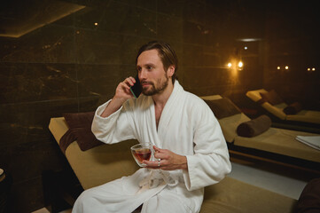 Relaxed handsome European man in white bathrobe holding a cup of tea and talking on cell phone,...