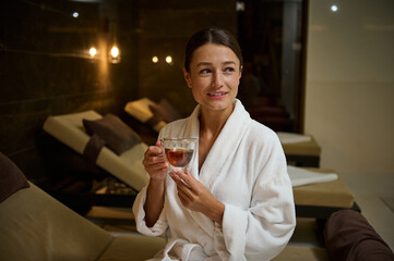 Beautiful elegant Caucasian young woman in white bathrobe, holding a glass cup of hot healthy herbal drink, looking aside, sitting on chaise lounge by jacuzzi, enjoying spa procedures in spa salon