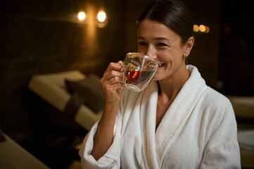 Charming elegant middle aged European woman in white bathrobe, drinking hot healthy herbal drink, sitting on chaise lounge by jacuzzi, enjoying spa procedures in spa salon