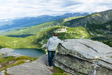 At the top of the high mountains you can see the mountains are covered with greenery and you can see the lakes from the melted snow. A man stands by a huge stone at the edge of the mountain top