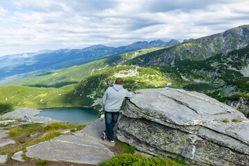 At the top of the high mountains you can see the mountains are covered with greenery and you can see the lakes from the melted snow. A man stands by a huge stone at the edge of the mountain top