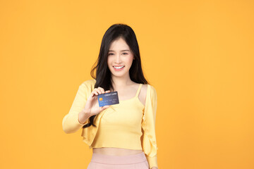 Cheerful young asian woman in yellow T-shirt holding bank card isolated on yellow background