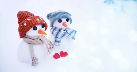 Two little cute snowmen in hats and scarves with red hearts in a snow-covered field close-up. Christmas background banner with winter symbols, romantic card for Valentine's day.	
