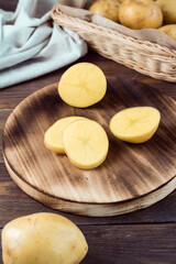 Fresh yellow potatoes cut into pieces on a board and a basket with tubers on a wooden table. Vegetarian food. Vertical view. Close-up