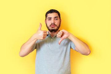 Confused bearded man is showing both like and dislike gestures over yellow background. Undecided...