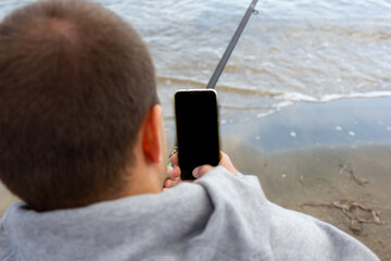 The phone in the hands of a man, view from the back. A man sits on a sandy shore near the water...