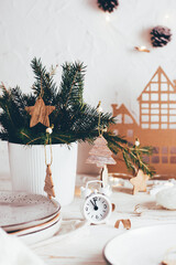 Organization of a festive Christmas table. White alarm clock, plates, spruce bouquet and Christmas decorations. Tablescapes on white wooden table. Vertical view