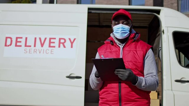 Portrait of african american courier delivering parcel box to recipient. Delivery man standing in front of a van wearing face mask, writting on tablet folder.