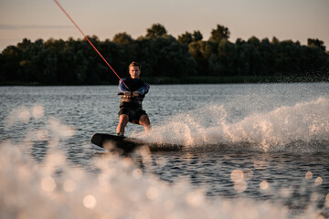 Fototapeta premium Great view of attractive man holding rope and energetically riding wakeboard on splashing river wave.