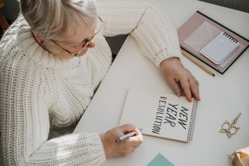 Healthy New Years resolutions for older adults. Senior mature old woman in white sweater writing handwritten text New Years resolutions in open notepad on the table. Planning and setting goals