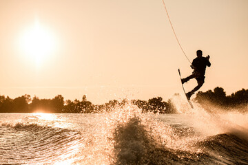 silhouette of male rider holding rope and making extreme jump on wakeboard over splashing water.