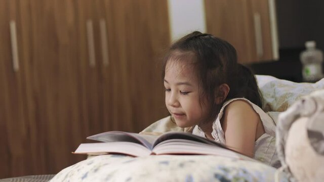 Adorable 5 years old Asian girl is reading a book, and lying on a bed with beautiful yellow light from a lamp. It is bedtime story for learning, relaxing and developing creativity of child.