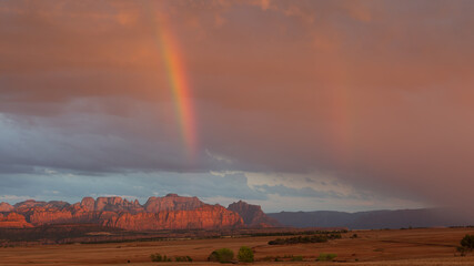 A rainbow arcs down towards the distant mountains of Zion National park at rain drifts down from passing storm clouds at sunset above Smiths Mesa in Southern Utah.