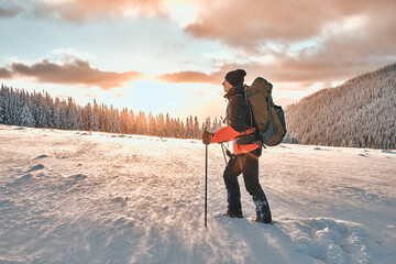 Handsome man dressed in winter sports warm clothes with a tourist backpack walks with trekking...