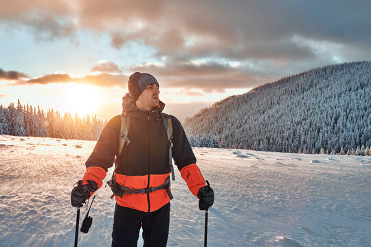 Handsome young man dressed in winter sports warm clothes stands with trekking poles and walkie-talkie in the snowy pine mountains at sunset. Copy space.