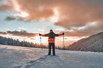 Happy man dressed in winter sports warm clothes with a tourist backpack and trekking poles in the...