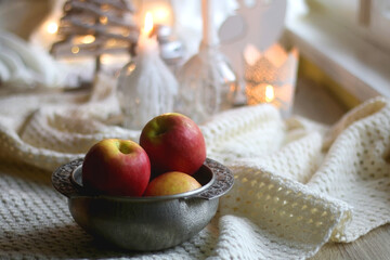 Bowl of apples and soft blanket with lit candles and Christmas decorations in the background. Selective focus.