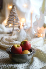 Bowl of apples and soft blanket with lit candles and Christmas decorations in the background. Selective focus.