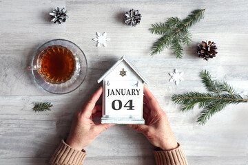 Calendar for January 4: hands hold a decorative house with the name of the month and the numbers 0 and 4, a cup with hot tea, snowflakes, fir branches on a gray background, top view