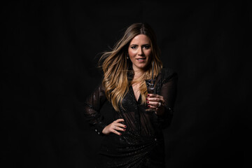 Attractive blonde woman in black sequin and glitter dress with glass in hand looking at camera and smiling on black background in photo studio