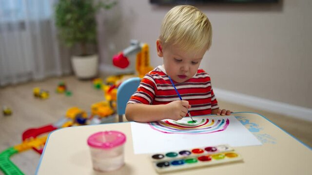 little boy with blond hair sits at the table and paints a rainbow.