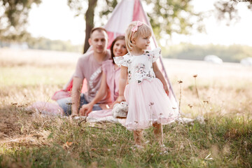 Mom, dad and little daughter are sitting next to wigwam decoration in the park. Family spending time outdoor in summer, having fun together. Girl are dressed in pink dress