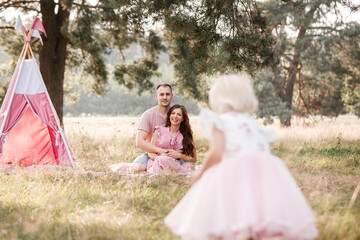 Mom, dad and little daughter are sitting next to wigwam decoration in the park. Family spending time outdoor in summer, having fun together. Girl are dressed in pink dress. © Andriy Medvediuk