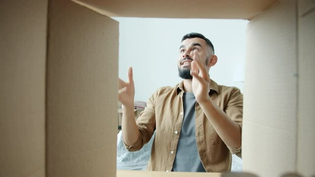 Cheerful Middle Eastern guy is unpacking things ball and books from cardboard box in new house smiling enjoying relocation. Youth and lifestyle concept.