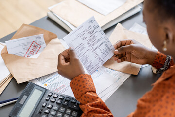 Over shoulder view of blackwoman sitting at table with bills and calculator and reading receipt